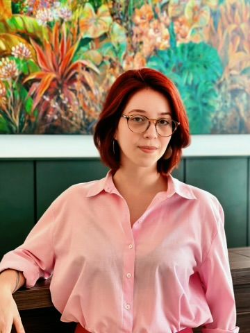 Nadyne, em camisa rosa, diante de um painel pintado com flora tropical.