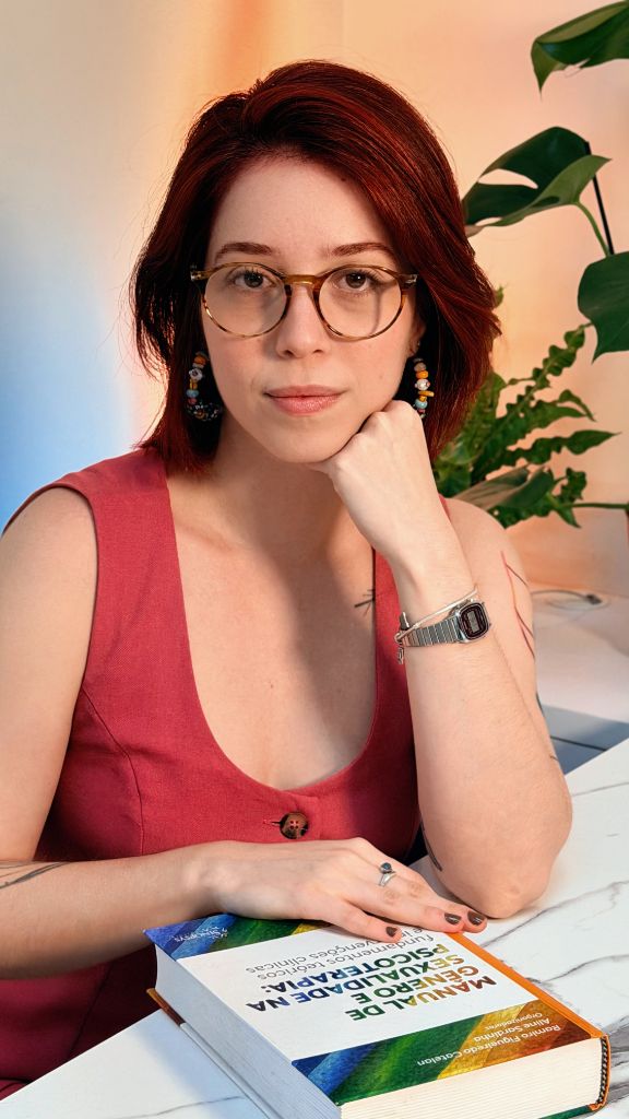 Nadyne, em retrato contemplativo, apoiada sobre um livro de psicoterapia, com folhagem ao fundo.